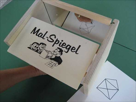 Pappkarton mit "Mal Spiegel" und Cartoon-Illustration, darunter eine Hand, die ein Blatt Papier hält.