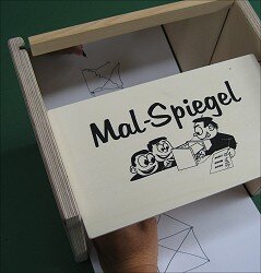 Holzbox mit Papier, auf dem "Mal-Spiegel" und eine Zeichnung eines Kindes mit Malutensilien.