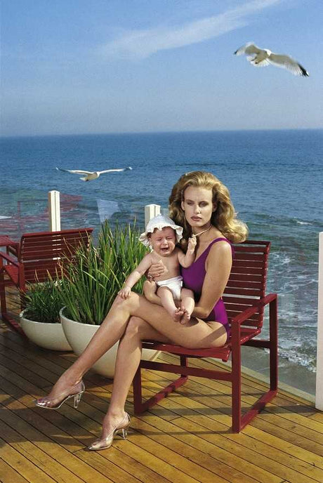 Schwangere Frau sitzt auf einer Bank am Meer, hält weinendes Baby, im Hintergrund fliegen Möwen.