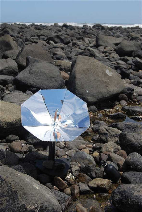 Ein reflektiernder Sonnenschirm auf einem felsigen Strand mit Wasser und Meer im Hintergrund.