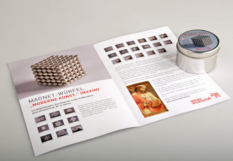 Offener Flyer mit Bildern und Text über Magnet-Würfel, daneben runde Metalldose mit Magnet-Würfel.