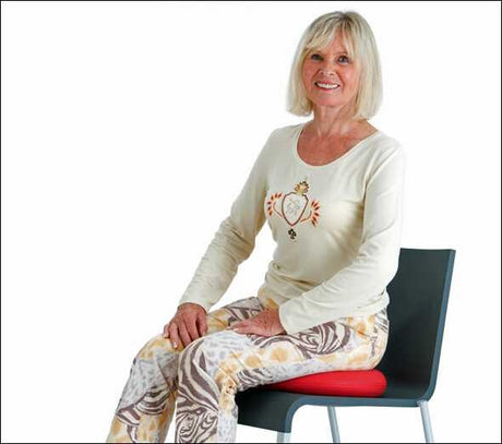 Ältere Frau mit blonden Haaren sitzt auf einem Stuhl mit roter Sitzfläche, trägt ein weißes Oberteil mit Hühner-Design.
