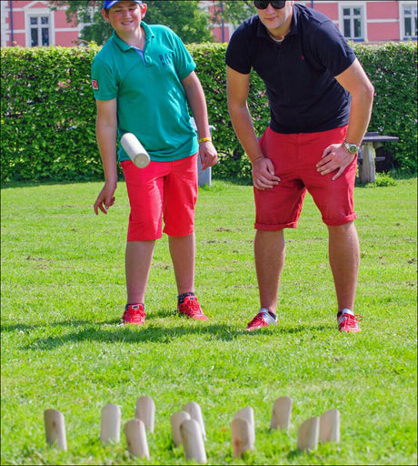 Zwei Personen beim Kubb-Spiel im Garten, einer wirft einen Holzklotz, die andere beobachtet.