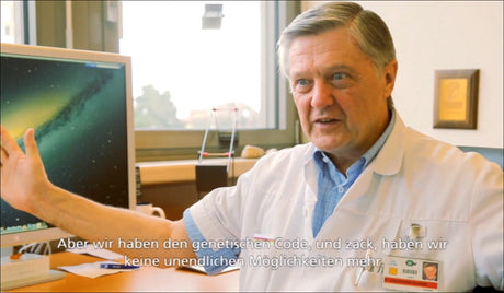 Älterer Mann in Arztkittel erklärt etwas vor einem Computerbildschirm, im Hintergrund Fenster und medizinische Utensilien.