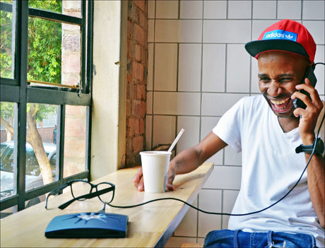 Junger Mann mit rotem Cap, weißem T-Shirt, spricht lachend am Telefon, sitzt an Tisch mit Brille, Laptop, Getränk und Sonnenbrille