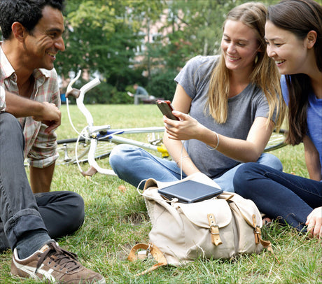 Drei junge Frauen und ein Mann sitzen im Park, lachen und schauen auf ein Smartphone. Fahrrad liegt im Hintergrund.