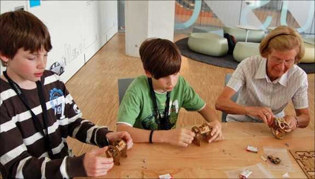 Zwei Jungen und eine ältere Frau sitzen an einem Tisch und bauen aus Holz kleine Figuren.