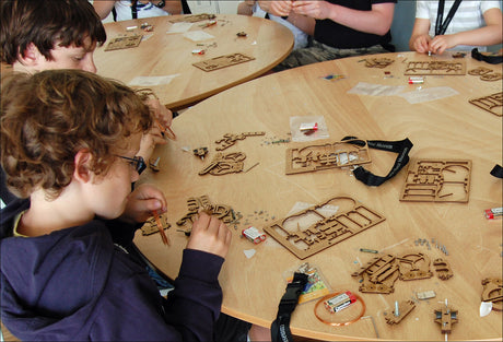 Kinder bauen mit Holzbausätzen an einem runden Tisch, umgeben von Werkzeugen und Batterien.