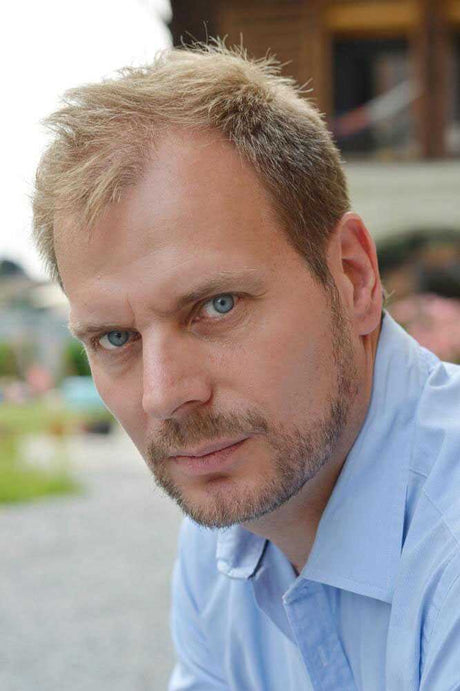 Nahaufnahme eines Mannes mit blauen Augen, kurzen hellen Haaren und Bart, trägt ein hellblaues Hemd.
