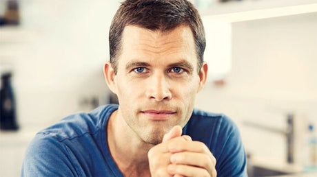 Junger Mann mit dunklem Haar und blauen Augen, trägt ein blaues T-Shirt, sitzt in einem Raum mit weißen Regalen im Hintergrund.