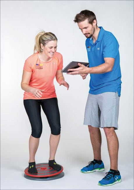 Frau steht auf einem Balance-Board, Trainer zeigt ihr etwas auf einem Tablet.