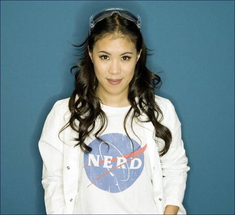 Junge Frau mit dunklen, lockigen Haaren, Brille auf Kopf, trägt weißes T-Shirt mit NASA-Logo und weißen Jacke vor blauer Wand.