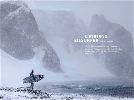 Ein einsamer Surfer mit Surfbrett steht im Schnee an einem eisigen Strand, im Hintergrund hohe, verschneite Felsen.