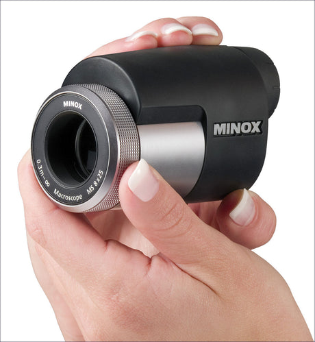 Hand hält Minox Mikroskop mit schwarzem Gehäuse und silbernem Objektiv, gegen weißen Hintergrund.
