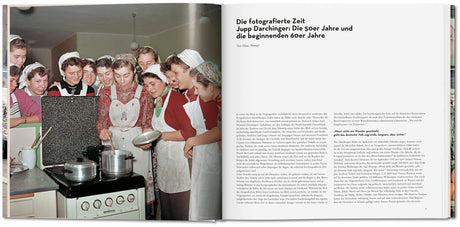 Mehrere junge Frauen in Küchenkleidung und Hauben schauen auf einen Herd mit Töpfen.