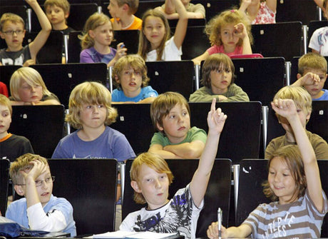 Kinder sitzen in einem Klassenraum, einige heben die Hand, andere schauen aufmerksam.