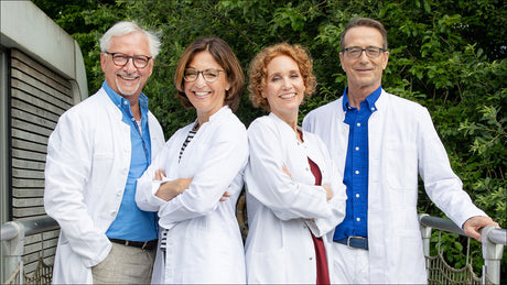 Vier medizinische Fachkräfte in weißen Kitteln stehen vor grünem Hintergrund, lächeln in die Kamera.
