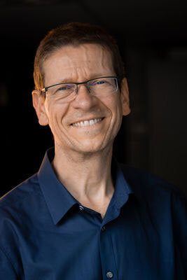 Lächelnder Mann mit Brille und dunklem Hemd vor dunklem Hintergrund.