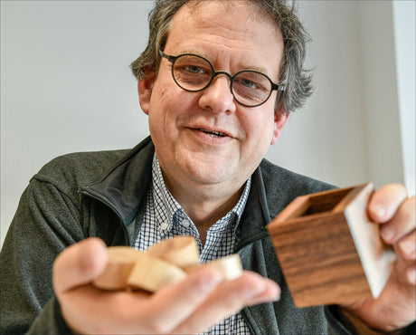 Älterer Mann mit Brille, lächelnd, hält Holz- und Holzoptik-Objekte in den Händen.
