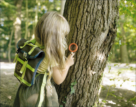 Mädchen mit Rucksack beobachtet Baum durch eine Lupe im Wald.