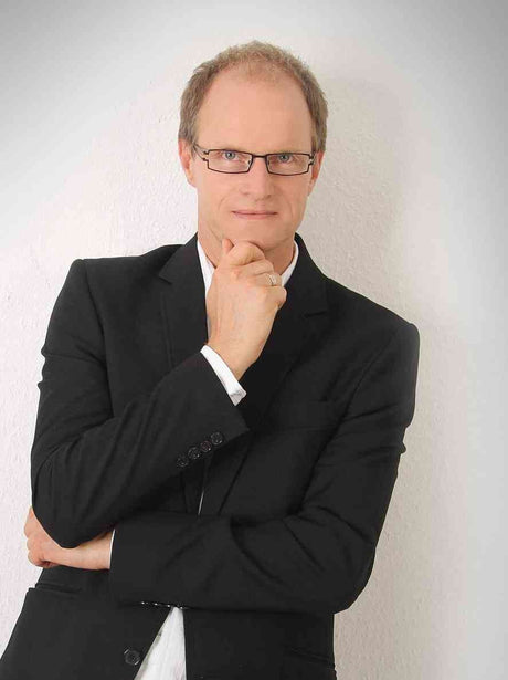 Mann mit Brille, schwarzem Anzug und weißem Hemd, nachdenklich mit Hand am Kinn vor weißer Wand.