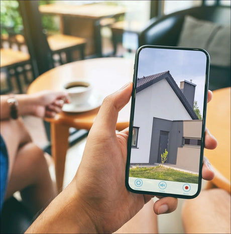 Hand hält Smartphone mit Bild eines Hauses, im Hintergrund Kaffee auf Tisch, Person mit Arm und Uhr.