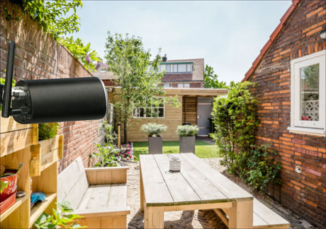 WLAN-Antenne an einer roten Backsteinmauer, Blick auf einen Garten mit Tisch, Bänken, Pflanzen und einem kleinen Holzhaus.