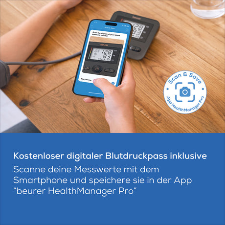 Hand hält Smartphone, zeigt Blutdruckmessgerät, im Hintergrund weiteres Blutdruckgerät auf Holz.