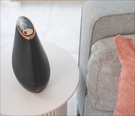 Luftbefeuchter auf einem weißen Tisch neben einem grauen Sofa mit rotem Kissen.