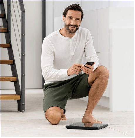 Mann kniet auf Waage, hält Smartphone, lächelt, trägt bequeme Kleidung, im modernen Zuhause.