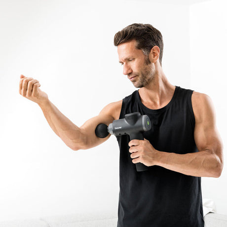 Mann mit Muskelzuwachs, der eine Massagepistole auf den Arm richtet, trägt schwarzes Tanktop, steht vor weißem Hintergrund.