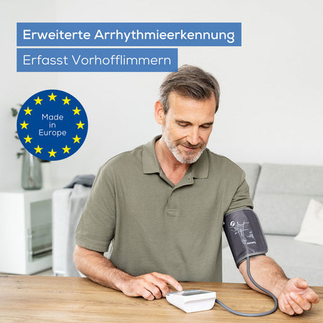 Älterer Mann misst Blutdruck mit Blutdruckmessgerät, sitzt an Tisch, trägt Hemd, im Hintergrund Sofa und Pflanze.