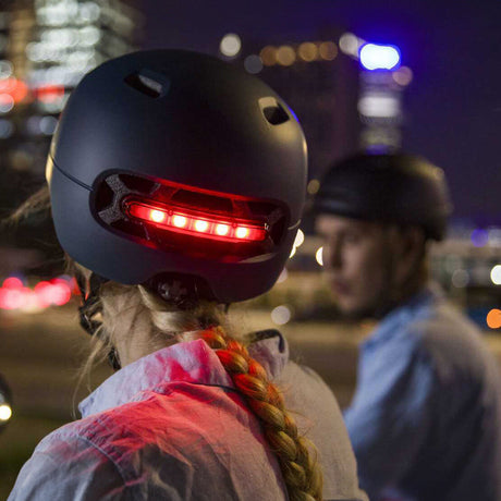 Zwei Personen mit schwarzen Helmen, eine mit geflochtener Haarpracht, bei Nacht, mit Stadtlichtern im Hintergrund.