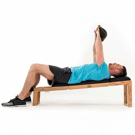 Mann liegt auf einer Bank, hebt mit beiden Armen eine schwarze Kettlebell.