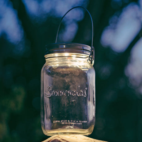 Glas mit Henkel, beschriftet mit "Sonnenglas", hängt an einem Draht vor dunklem, unscharfem Hintergrund.