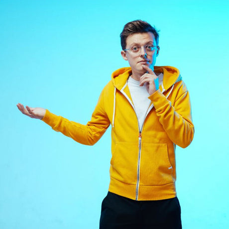 Junger Mann in gelber Kapuzenjacke, trägt Brille, steht vor blauem Hintergrund, wirkt nachdenklich, mit einer Hand gestikuliert.