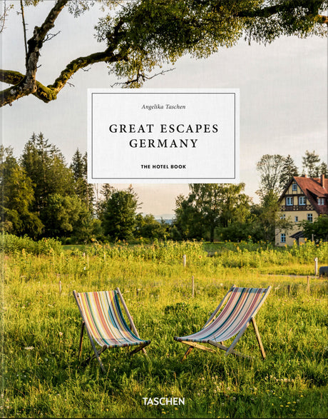 Zwei bunte Strandstühle auf einer grünen Wiese mit Bäumen und einem Haus im Hintergrund, bei Sonnenuntergang.